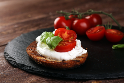 Na imagem, há uma torradinha com creme de ricota e duas rodelas de tomate