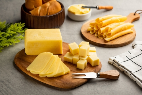 Na imagem, há uma tábua de madeira com queijo mussarela em peça, cubinhos e fatias